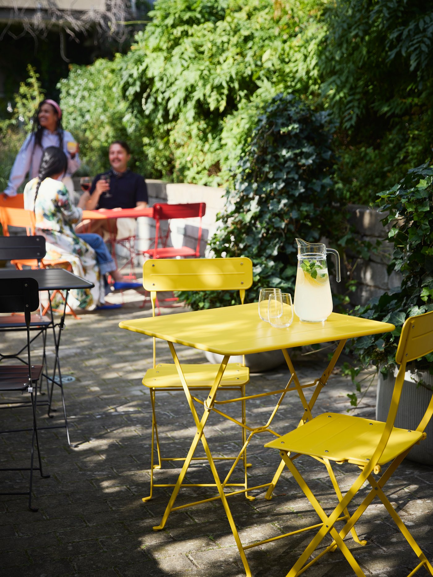 Bright SUNDSÖ tables in red, black, and yellow in a sunny courtyard, with people enjoying their time in the background.
