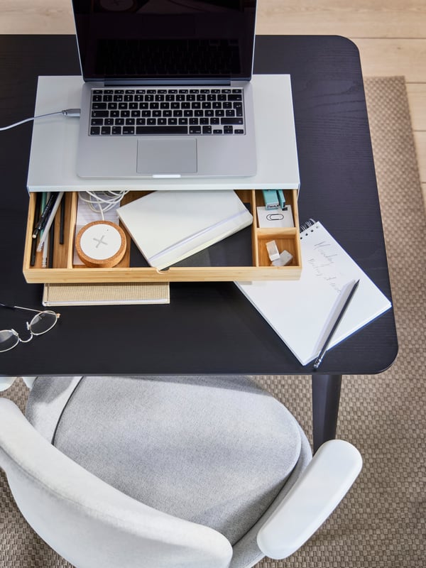 An ELLOVEN monitor stand on a LISABO dining table with a laptop on top and the drawer open showing a notebook and a charger.