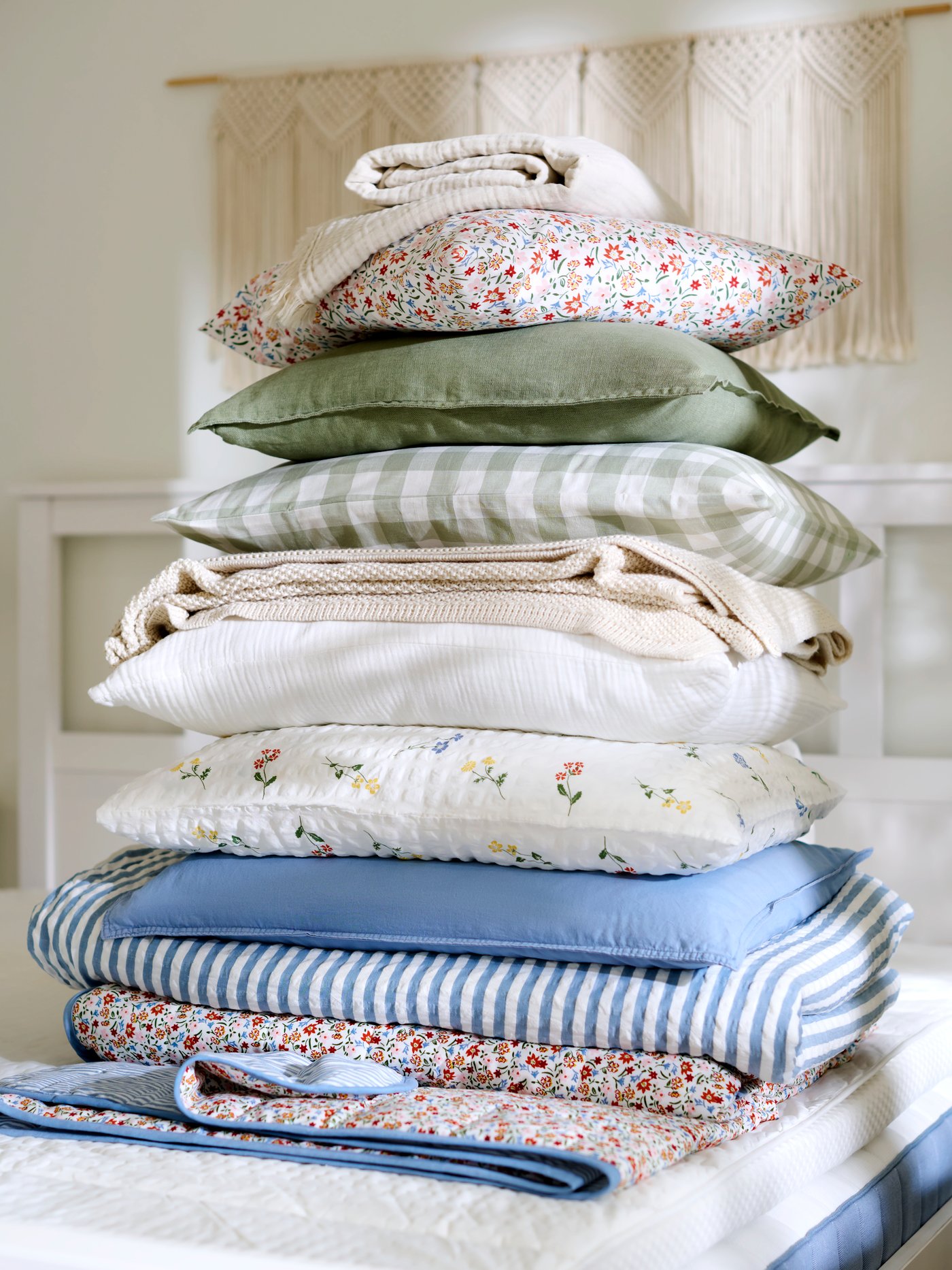 A big pile of pillows, duvets and comforters in light colours and summer patterns, on top of a bed.
