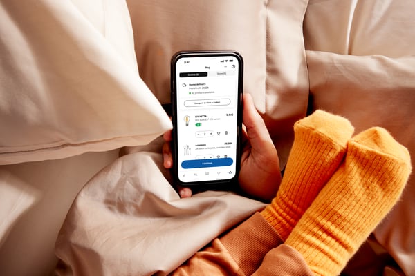 Na krevetu, nečija ruka drži pametni telefon na kom je prikazana IKEA aplikacija, a stopala malog deteta s narandžastim čarapama naslonjena su na ruku te osobe.