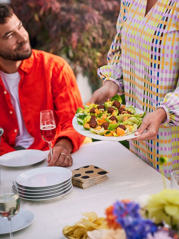 Iemand zit aan een tafel en ernaast staat iemand die een bord met HUVUDROLL plantaardige balletjes en salade vasthoudt.