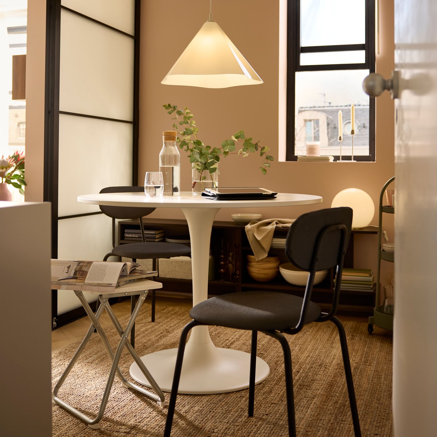 Black ÖSTANÖ chairs and a white GUNDE folding stool surrounding a white DOCKSTA table in a dining area with sliding doors.