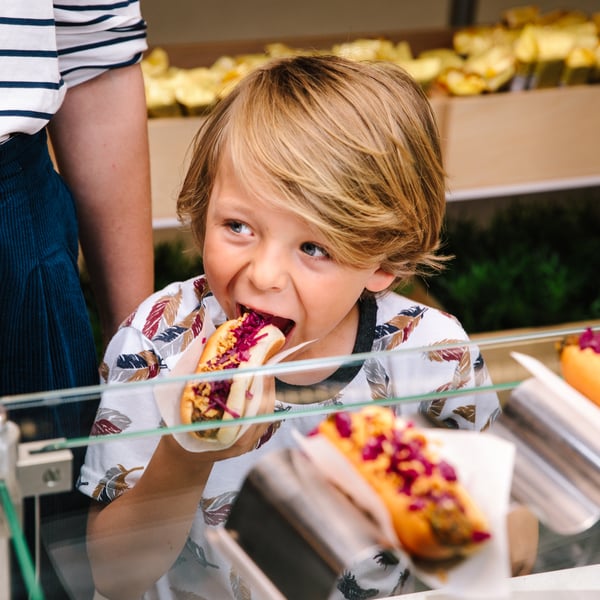 Enfant prenant un hot‑dog sur un comptoir de vente, entouré de garnitures colorées et d’ingrédients frais dans un environnement animé.