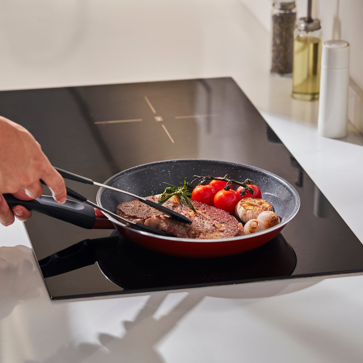 Hand uses tongs to cook steak with tomatoes and mushrooms in a skillet on an induction cooktop.