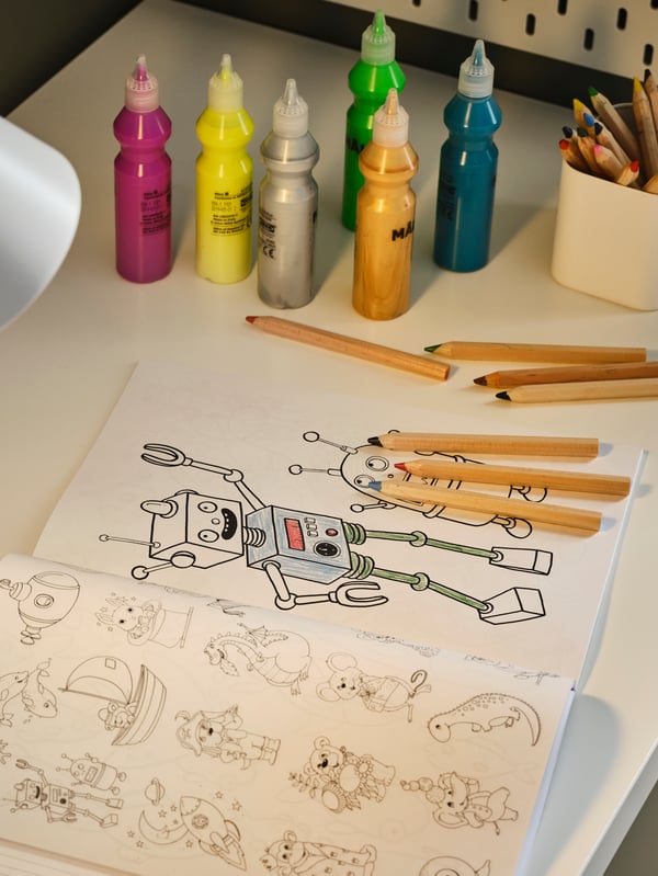 Un escritorio con un libro para colorear MÅLA abierto y parcialmente coloreado, pintura de purpurina MÅLA variada y lápices de colores MÅLA.