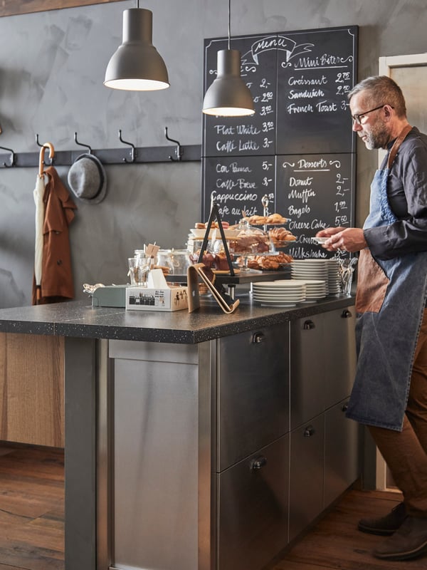 A man wearing an apron standing behind a black SÄLJAN worktop with mineral effect, with a hand-written menu behind.