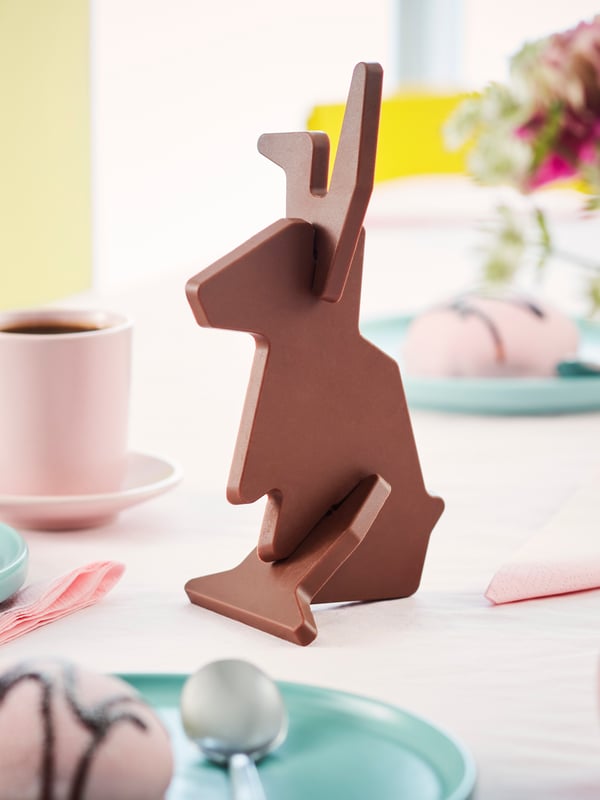 An assembled VÅRKÄNSLA milk choco bunny on a tablecloth next to a pink cup, turquoise plates with cake and pink napkins.