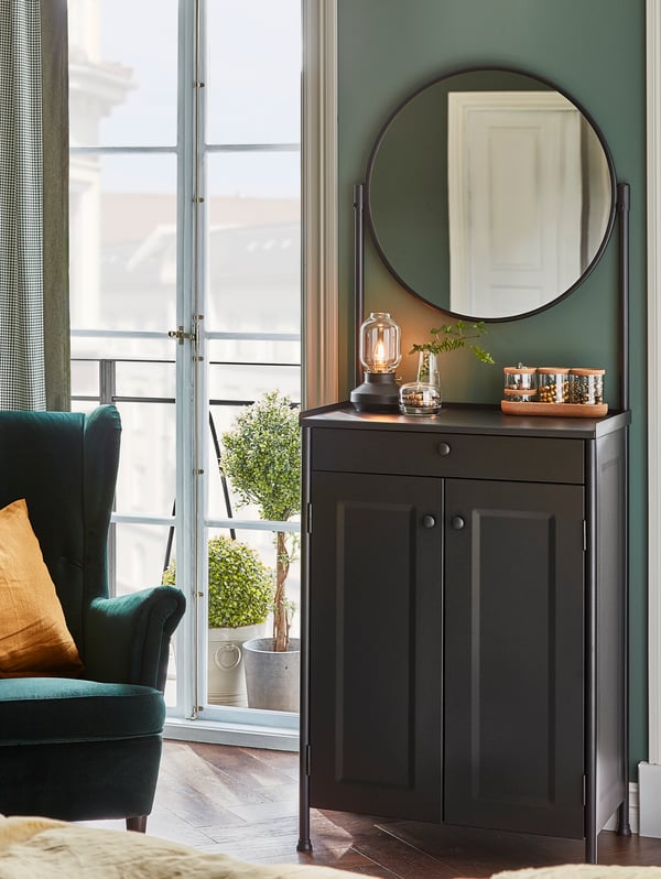 A KORNSJÖ cabinet with mirror stands against a wall. A table lamp, a vase and glass jars with lids stand on top of it.