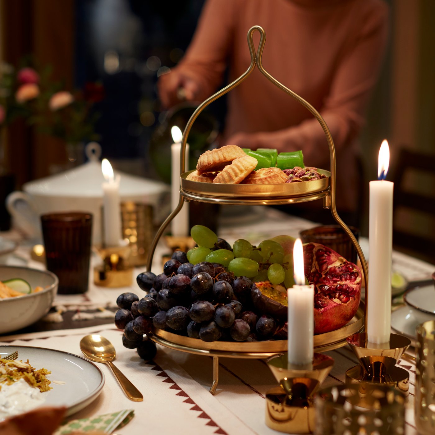 A golden GOKVÄLLÅ two-tier serving stand displays grapes, pomegranate, and biscuits on a decorated table with candles.