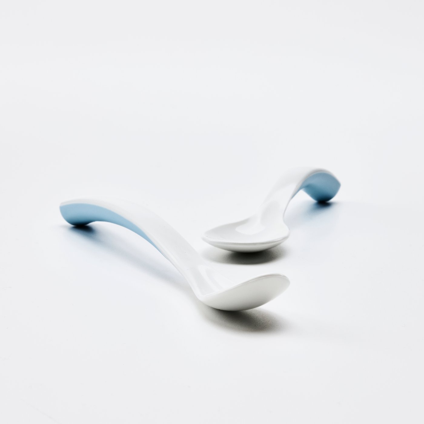 A blue/white BÖRJA feeding spoon and baby spoon sit on a white surface in a completely white environment.
