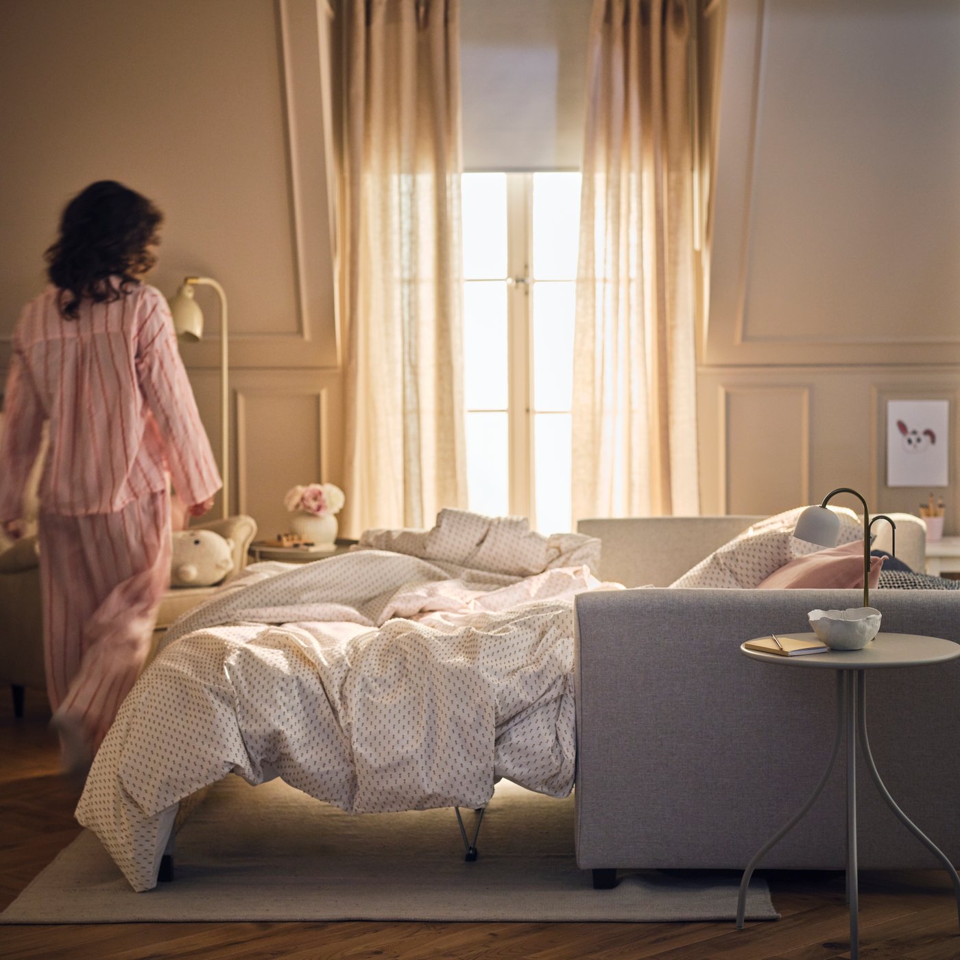 A person stands beside a light beige EKHOLMA 2-seat sofa-bed which is configured as a bed near a window with DYTÅG curtains.