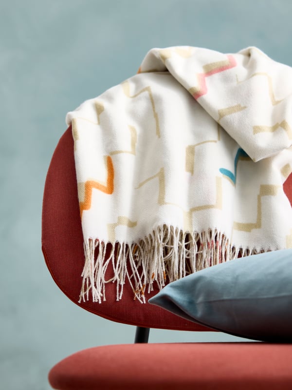 Close up of a OLIVSPINNMAL throw and a SANELA pillow placed on a red GÄVLE easy chair.