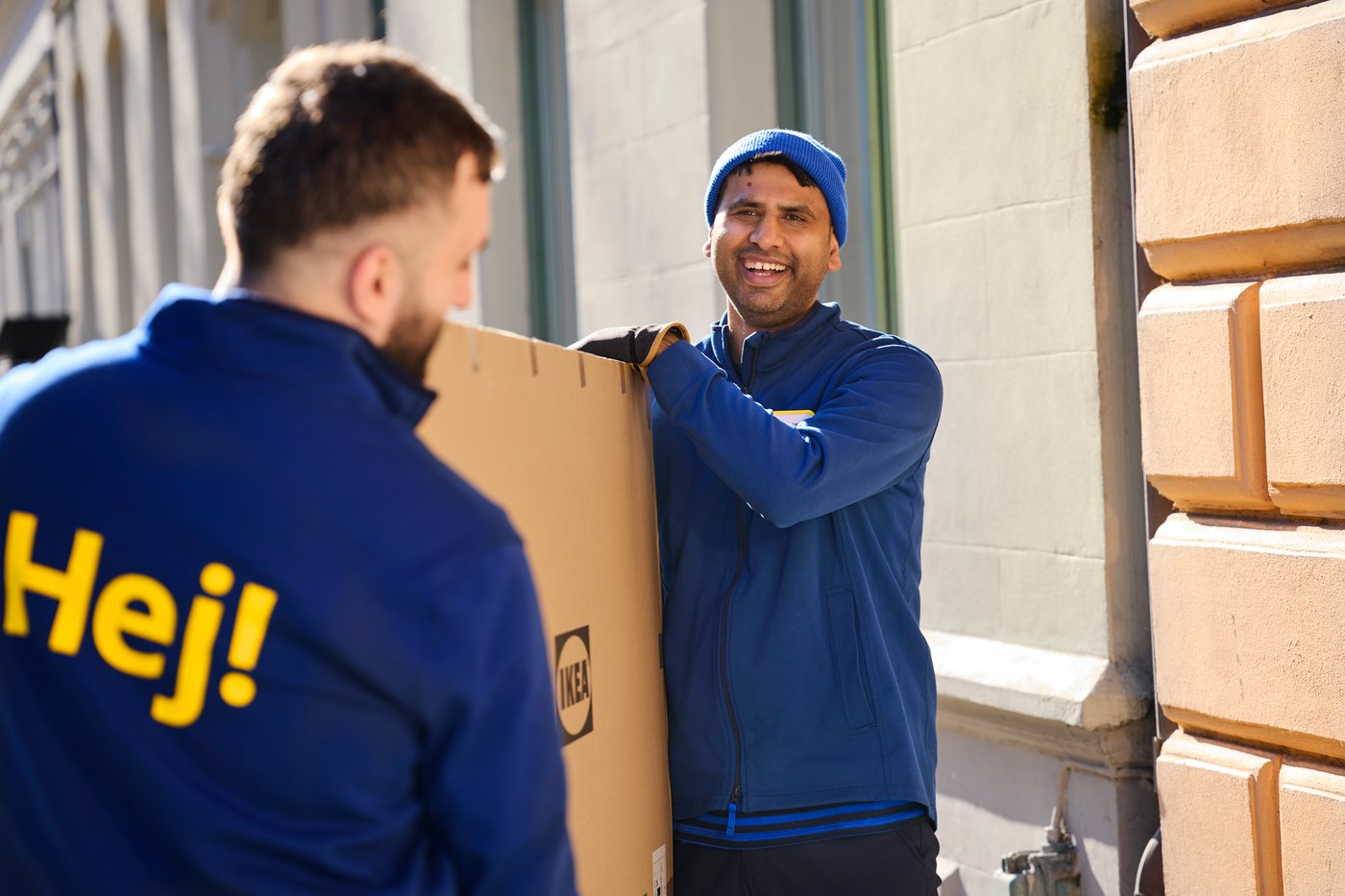 Two people in blue work clothes carry a large cardboard box together outdoors, with the text “Hello!” visible on a jacket.
