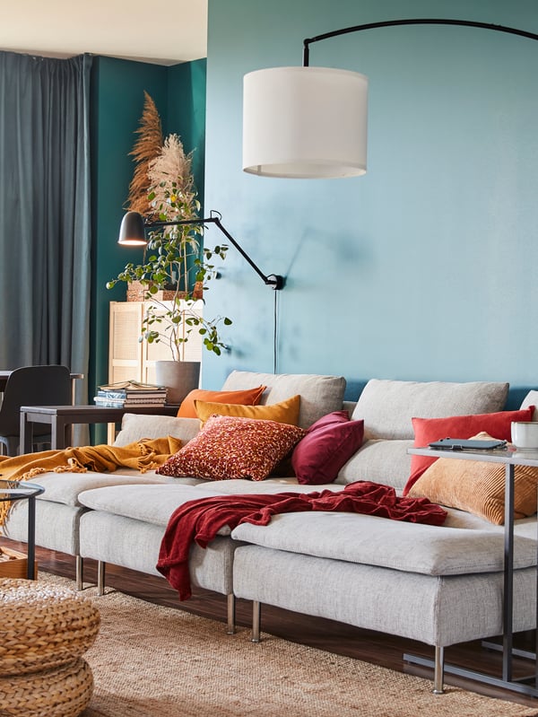 Drei SÖDERHAMN Récamieren mit Bezug „Viarp“ in Beige/Braun ergeben nebeneinander ein grosszügiges Sofa. Davor liegt ein Juteteppich, daneben steht eine Standleuchte in Bogenform und auf dem Sofa liegen viele Kissen in Rot/Gelb. 