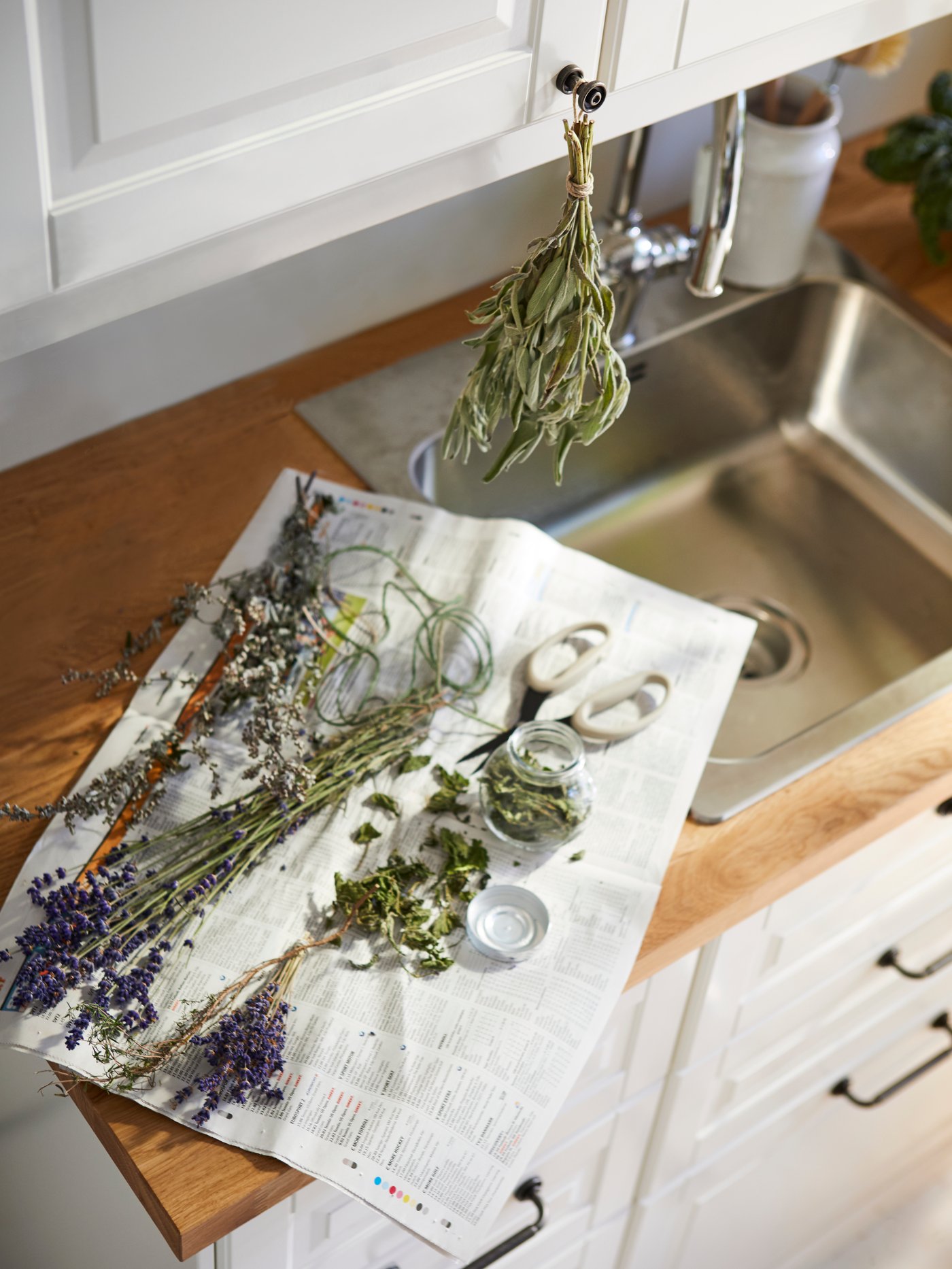 Un plan de travail de cuisine avec un évier est recouvert d’un journal sur lequel reposent diverses herbes et fleurs séchées, accompagnées d’un petit pot en verre, de ciseaux et de ficelle, tandis qu’un bouquet d’herbes séchées est suspendu à une poignée de placard au-dessus.