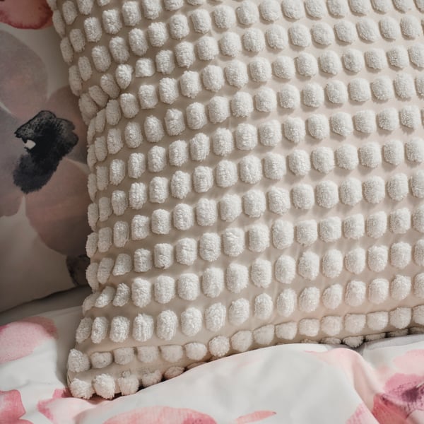 A cushion covered in an off-white SVARTPOPPEL cover on top of bedding in floral white/multicoloured LAPPNYCKLAR bed linen.