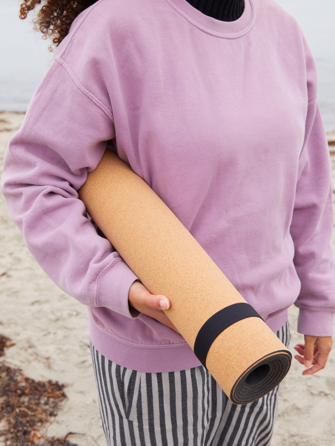 A woman wearing a purple sweatshirt holding the orange KÅSEBERGA yoga mat under her arm by a sandy beach by the sea.