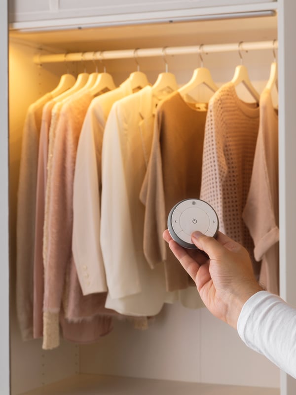 A hand holding a remote control, adjusting the smart lighting in a storage unit filled with clothes.