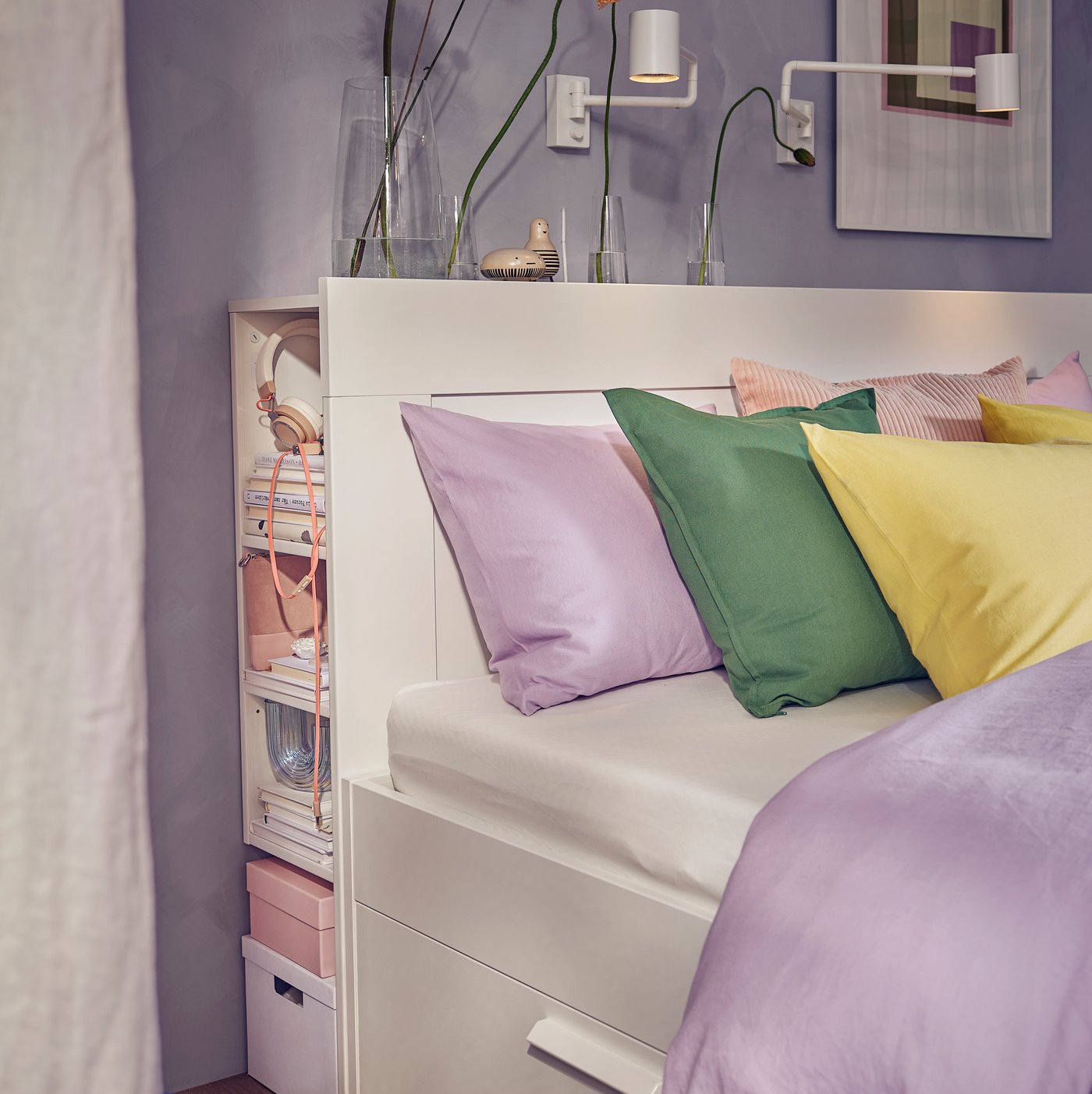 A BRIMNES bed with storage and headboard, with NÄTTSVÄRMARE bed linen and a green cushion, stands against a wall.