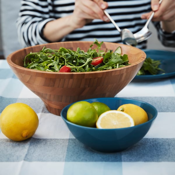 FARGKLAR bowl in blue filled with lemons, together with a GRONSAKER salad serving bowl with a salad inside, and a lady enjoying the salad at the dining table with a FARGKLAR blue plate and DRAGON cutleries. 