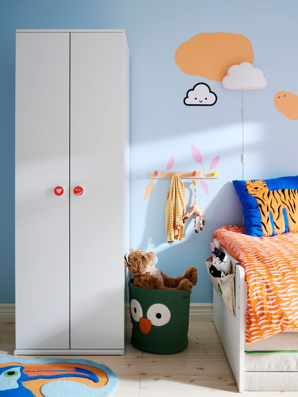 The foot-end of a SLÄKT bed by a light blue wall, with GODISHUS wardrobe to the left and wall-mounted hooks in between.