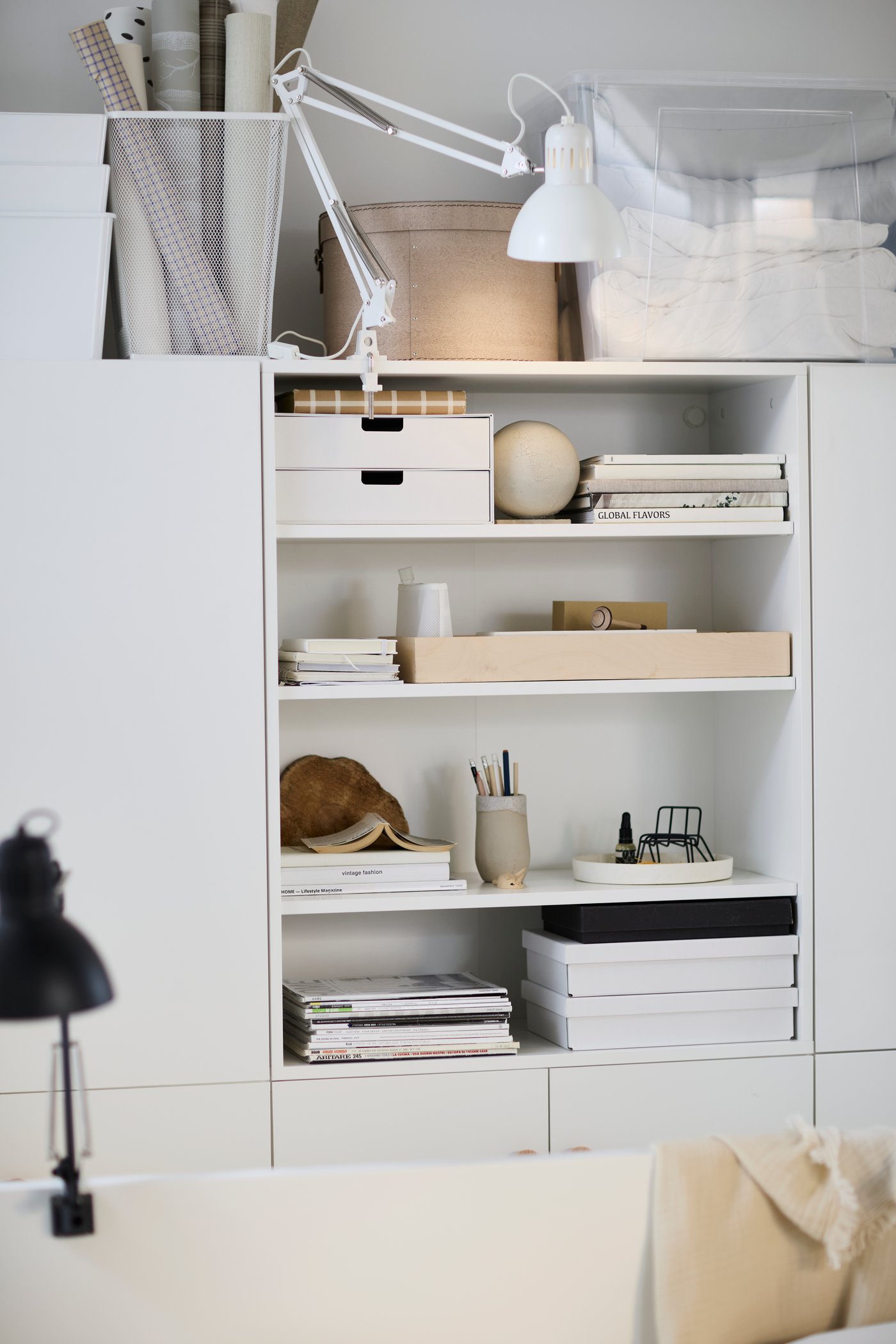 Vit hylla med öppna fack, fylld med böcker, förvaringslådor, skrivmaterial och en liten lampa; ovanpå hyllan står pappersrullar och en genomskinlig plastlåda.