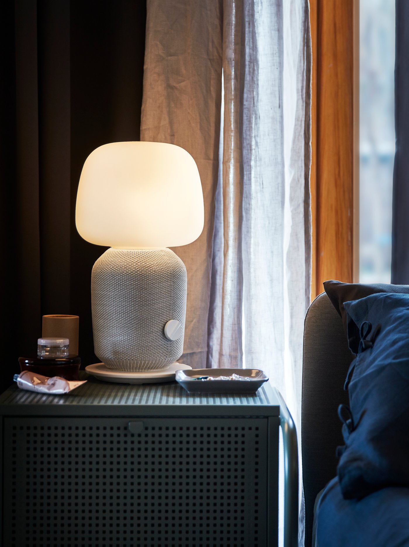 A night stand with a white and grey SYMFONISK table lamp with WiFi speaker standing in front of a window with grey curtains.