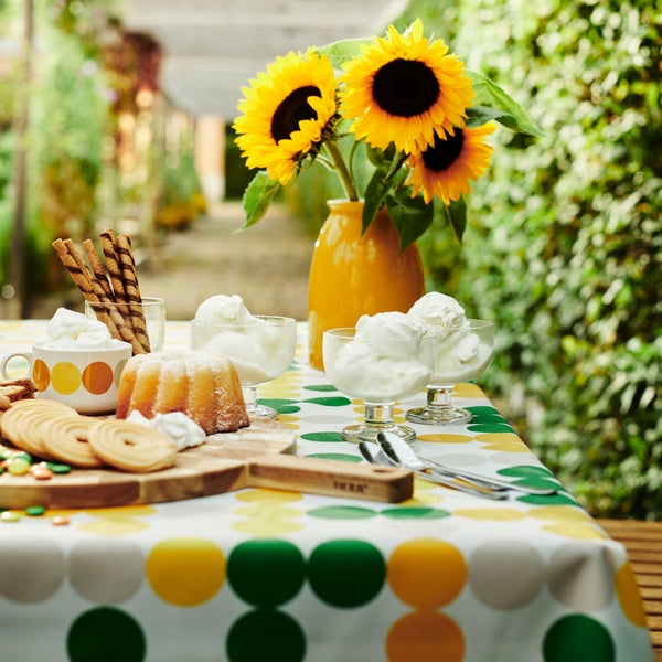 A table made with a dot-patterned BRÖGGAN tablecloth, tableware and a yellow vase with sunflowers, surrounded by greenery.