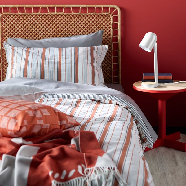 A desk lamp sitting on a red side table beside a bed with a rattan headboard, featuring a red-brown GULDAKLEJA comforter set.