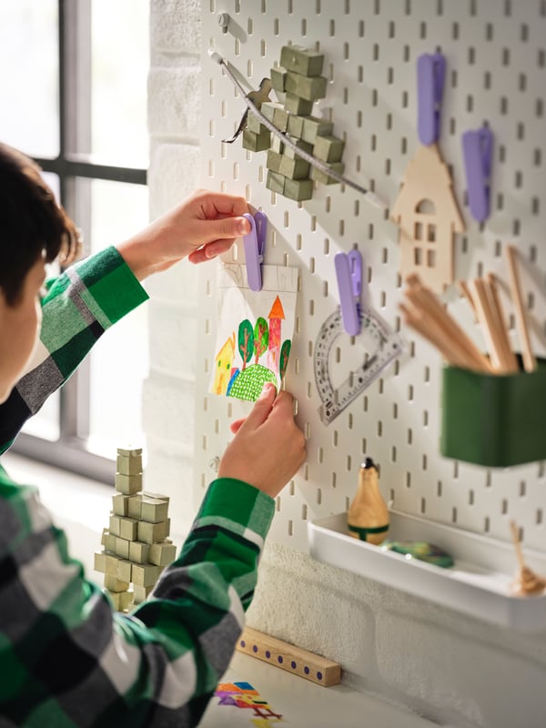 Close-up of tween boy putting pictures up on white pegboard using purple clips above a white children’s desk.
