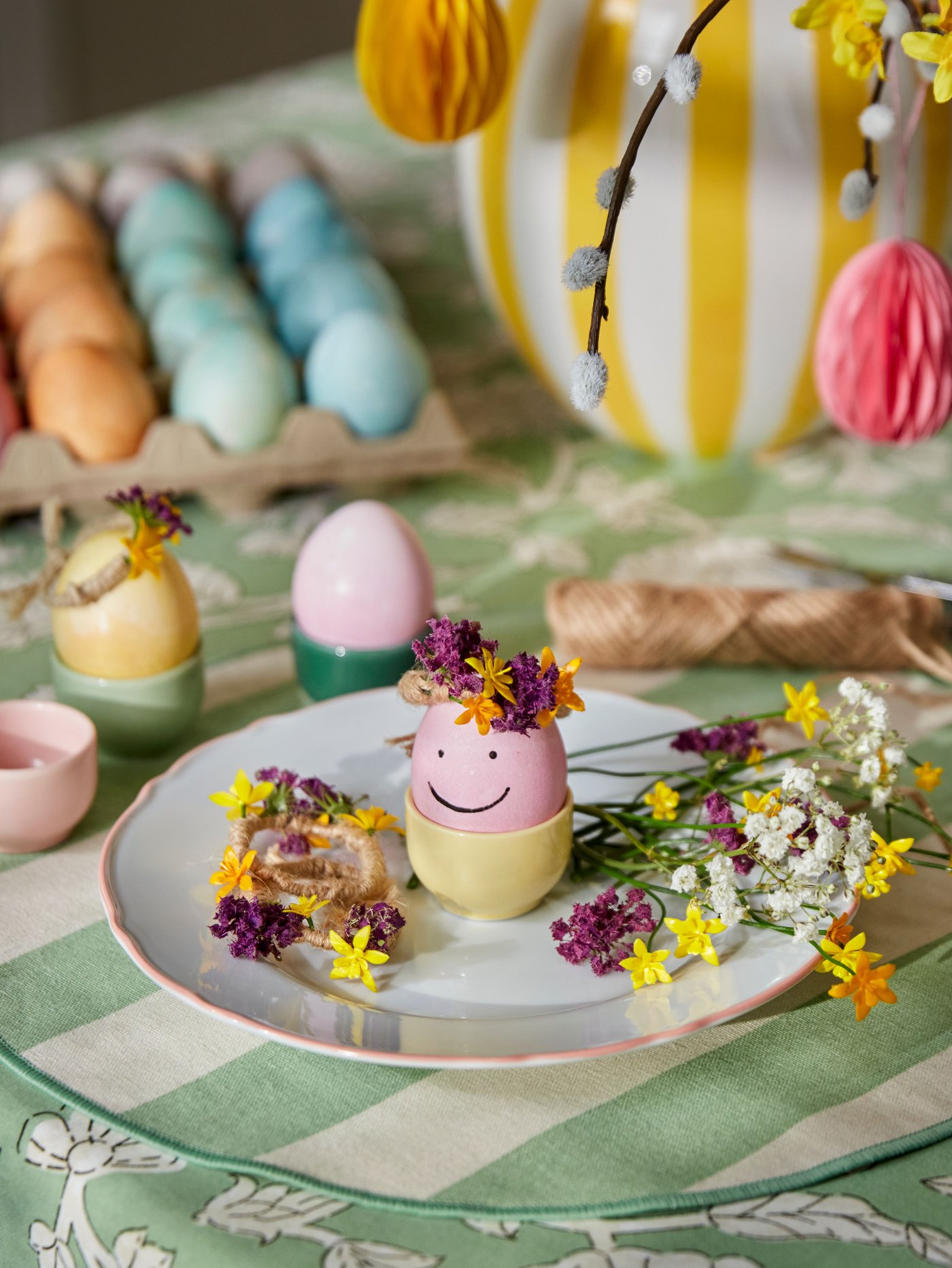 A plate with a colorful egg with a smiley face in an egg cup and flowers sits on a dining table. In the background, more colorful eggs and a white/yellow vase can be seen.