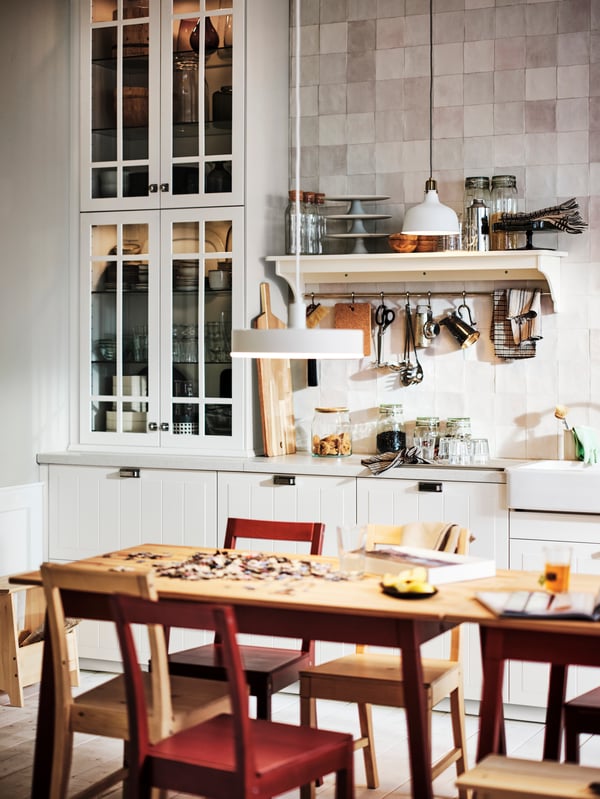 A kitchen with white STENSUND drawer fronts and glass doors and two PINNTORP tables surrounded by PINNTORP chairs.