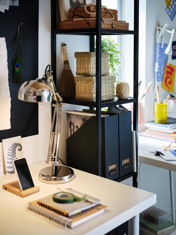 À côté du bureau avec un plateau LAGKAPTEN blanc éclairé par une lampe de bureau FORSÅ, il y a une structure murale haute cinq étagères ENHET.
