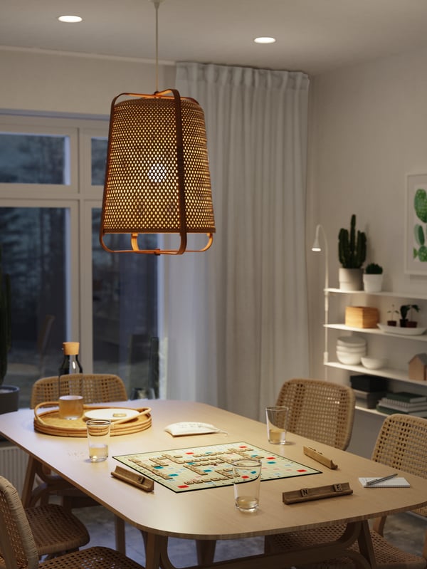 A dining room with bamboo and paper loom tables and chairs, a board game, a lit bamboo pendant lamp.