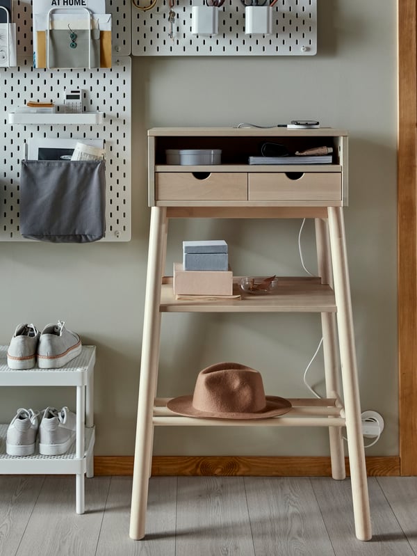 A KNOTTEN standing desk holding books, a charger and a hat, with a memo board above, by a MACKAPÄR shoe rack holding shoes.