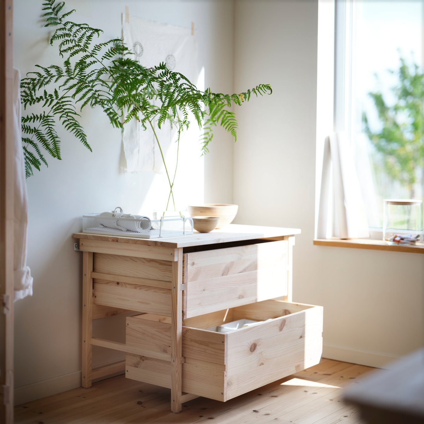 KRONÖREN kommode med 2 skuffer af fyrretræ med plante og skål ovenpå placeret ved siden af et vindue i et lyst soveværelse.