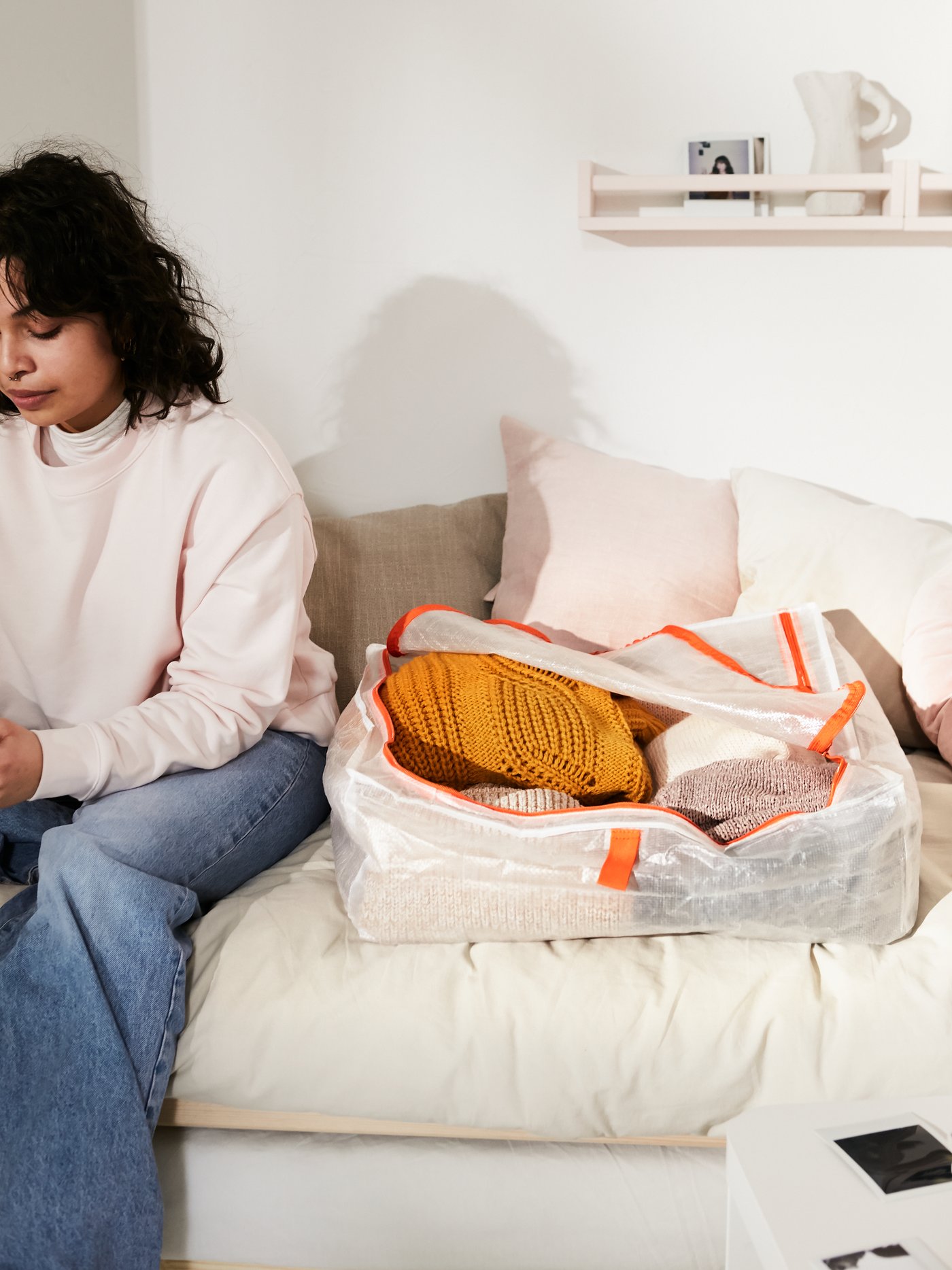 A young woman sitting on a bed next to a white PÄRKLA storage case with orange details, filled with clothes.