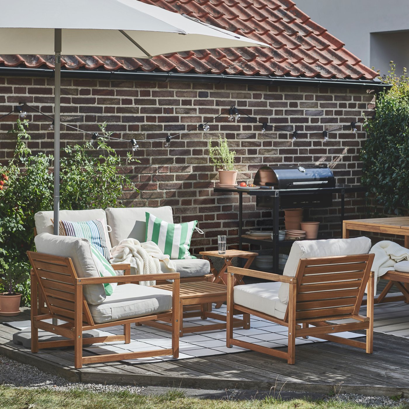 NÄMMARÖ wooden outdoor seating with cushions arranged under a parasol in a garden. 