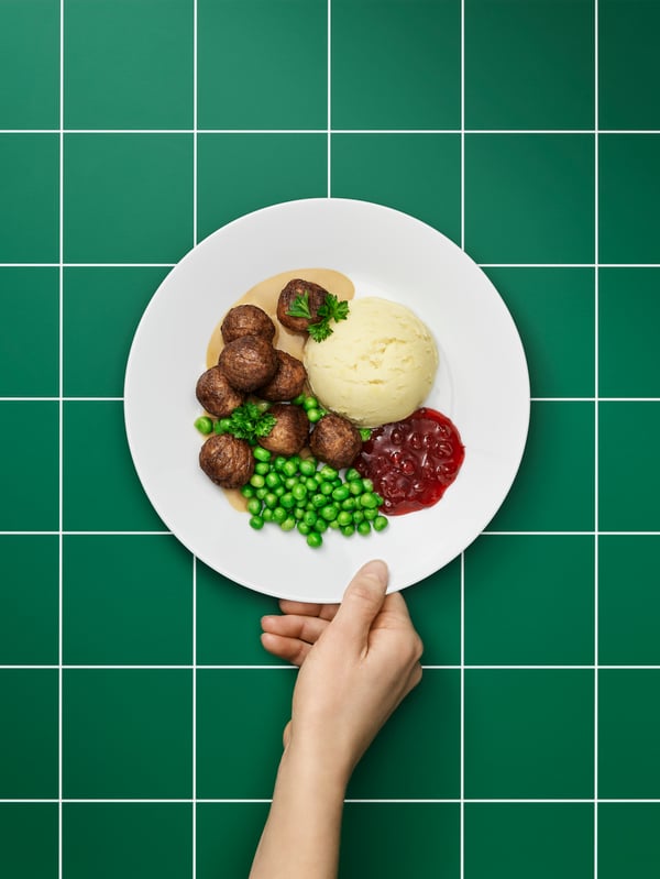 Une main tient une assiette remplie de boules de plantes HUVUDROLL sur un fond à carreaux verts et blancs.