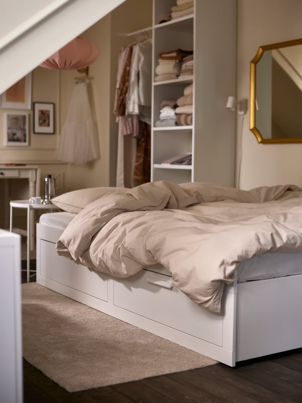 Un diván-cama BRIMNES blanco ampliado con ropa de cama gris claro/beige, con un estante blanco y un espejo con marco dorado