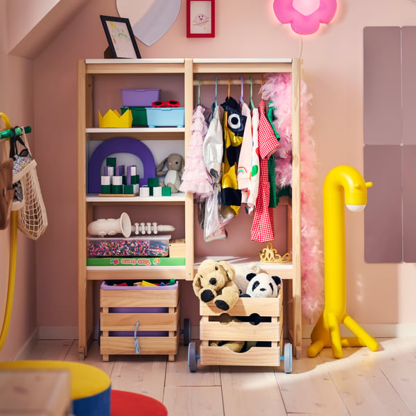 A pink children’s room with a FLISAT open storage with shelves, a clothes rail with dress-up clothes and wheeled boxes.