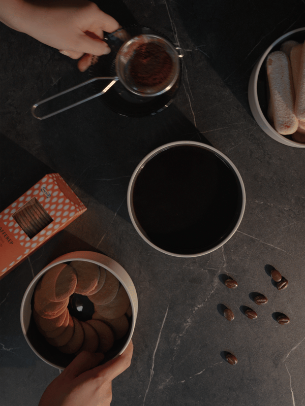 Hands prepare coffee using a strainer over a black cup on a dark surface, surrounded by a round tin of biscuits, sponge fingers and scattered coffee beans. The atmosphere is cosy and inviting.