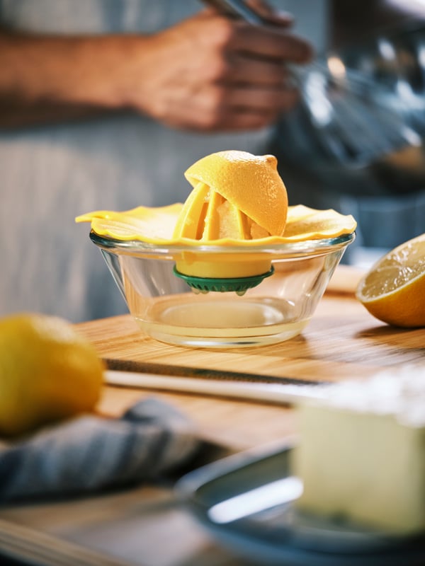 An APTITLIG chopping board in bamboo holding an UPPFYLLD lemon squeezer in bright yellow with the arm of a person behind.