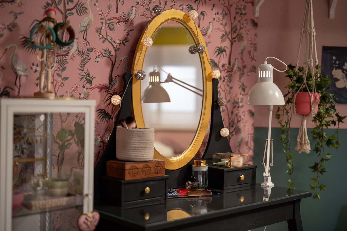 Een zwarte kaptafel met een ovaal goudkleurig HEMNES spiegel, gecombineerd met hanglampen en planten, staat tegen een roze bloemenmuur en groene wand