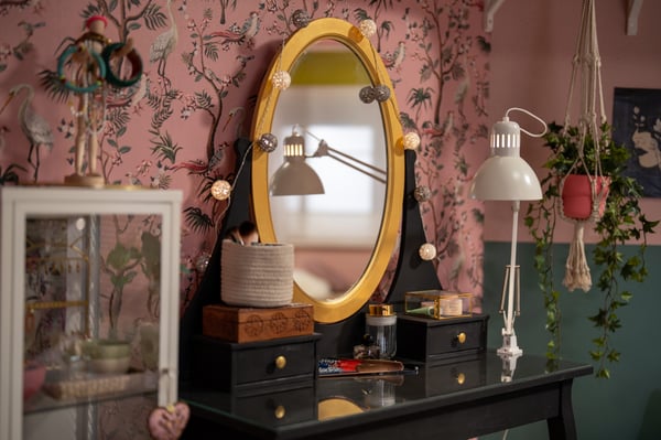 Een zwarte kaptafel met een ovaal goudkleurig HEMNES spiegel, gecombineerd met hanglampen en planten, staat tegen een roze bloemenmuur en groene wand