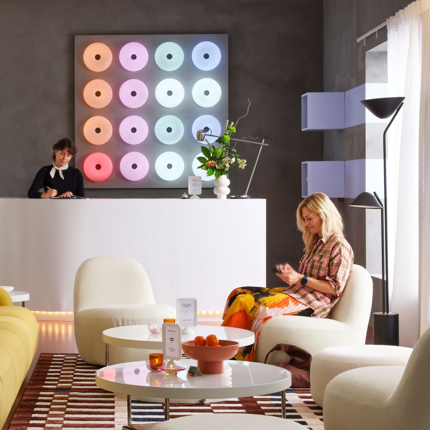 A square panel has 16 donut-shaped VARMBLIXT table/wall smart lamps in various colours behind an reception desk in a lounge.