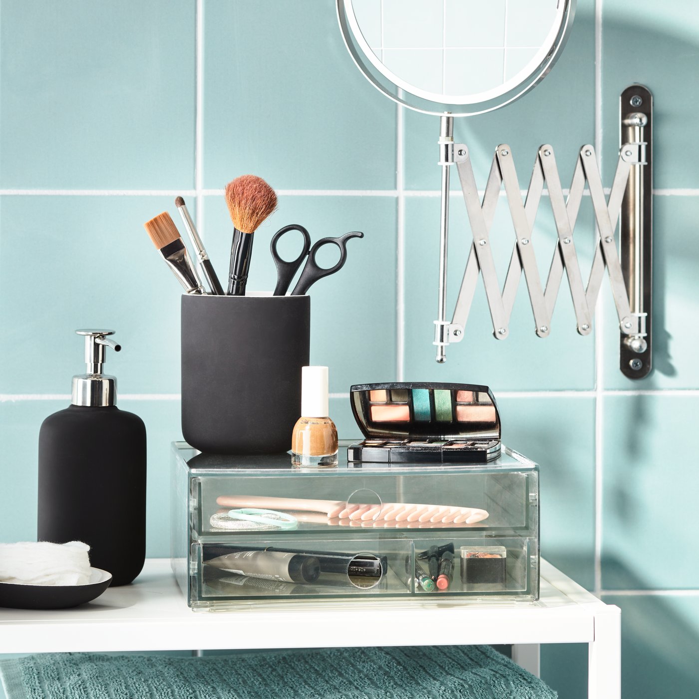 A stylish IKEA bathroom arrangement features a black soap dispenser, a makeup-brush holder, cosmetics, a magnifying mirror, and transparent storage drawers against glossy turquoise tiles.