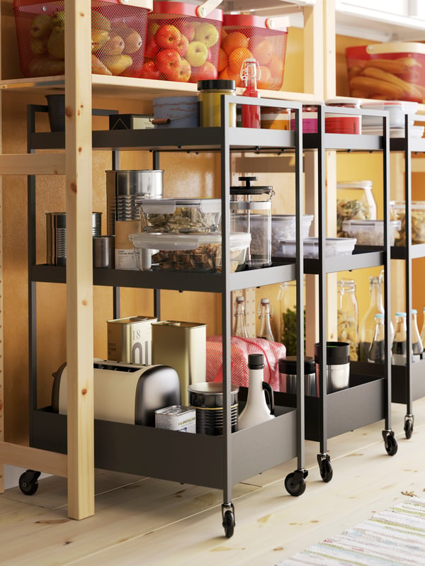 Three black trolleys with castors stand under a shelving unit in pine, and the trolleys store food and kitchen appliances.