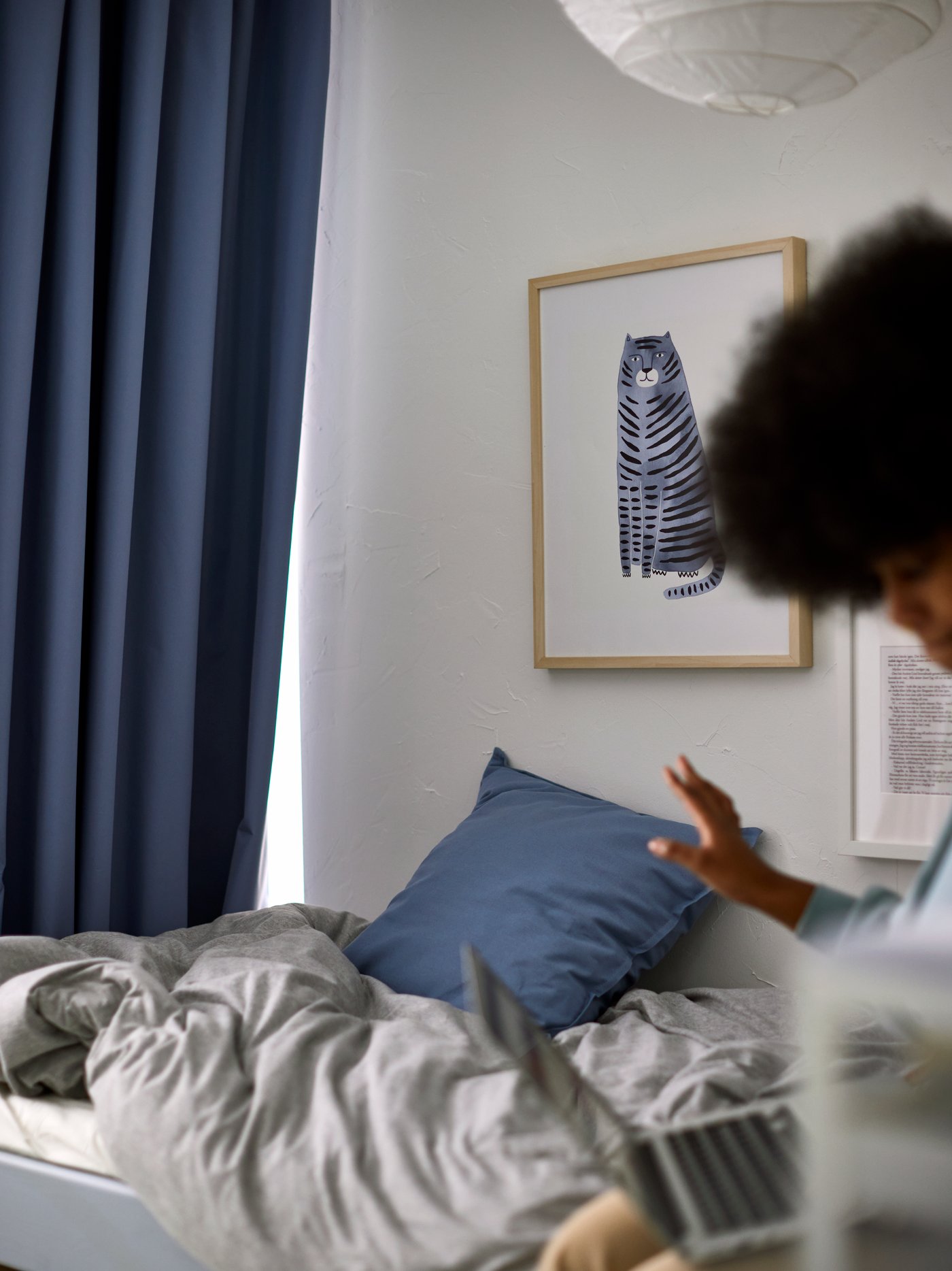 A woman with a computer on a bed with light gray bed linen, with a blue BENGTA block-out curtain by the foot of the bed.