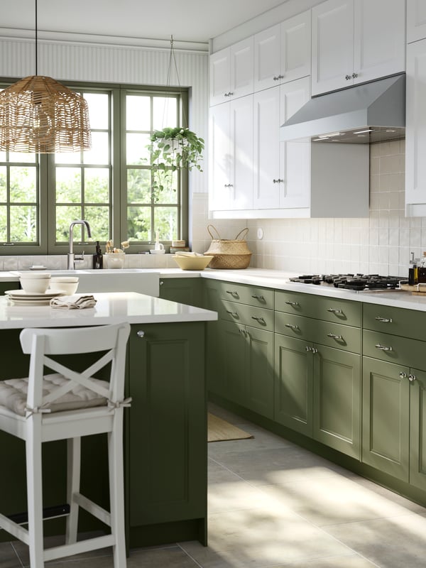 A kitchen with a large window has cabinets with grey-green and white AXSTAD fronts and a kitchen island with seating.